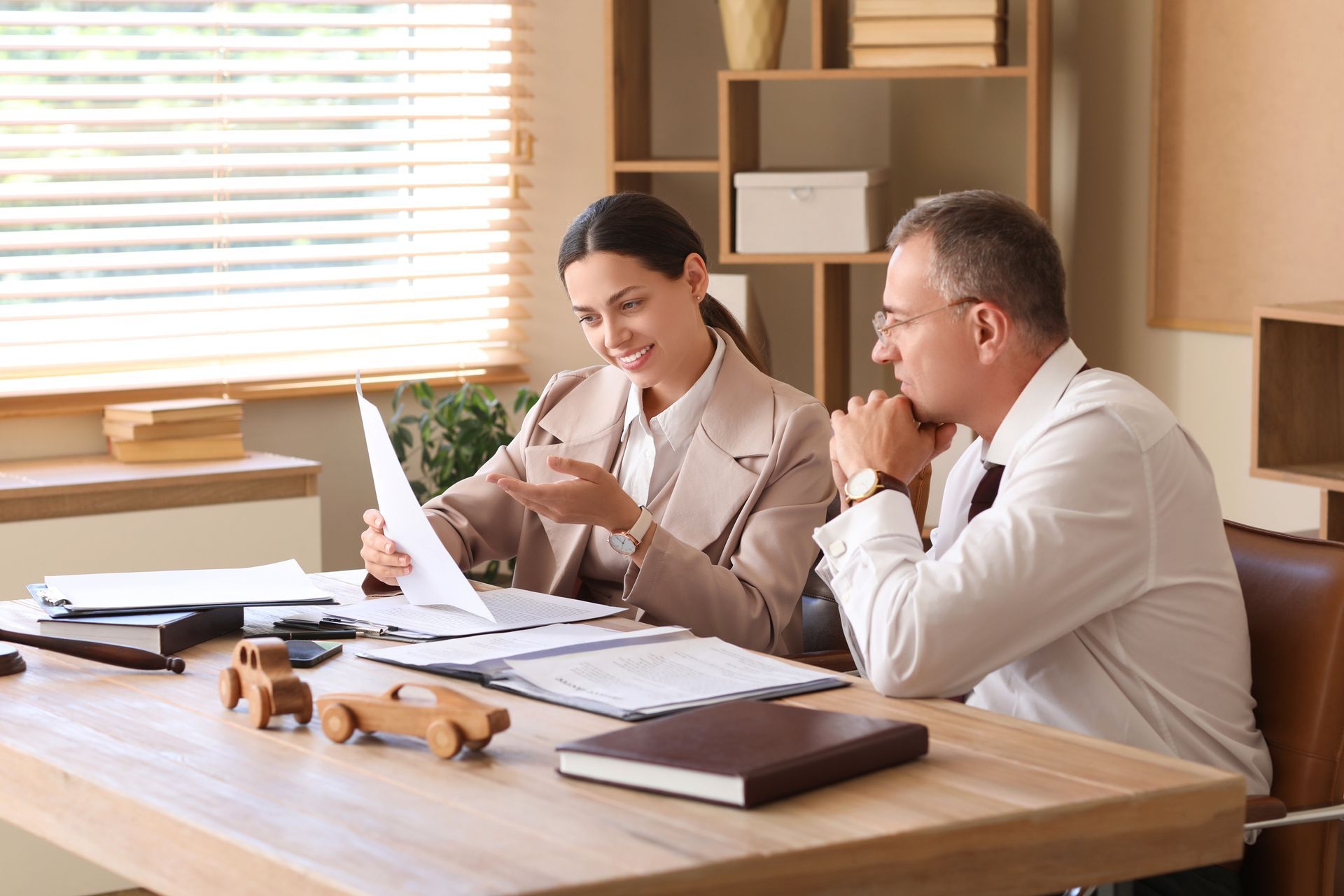 Woman and man reviewing documents at a desk in an office. Wooden cars on the desk. Woman and man reviewing documents at a desk in an office. Wooden cars on the desk.