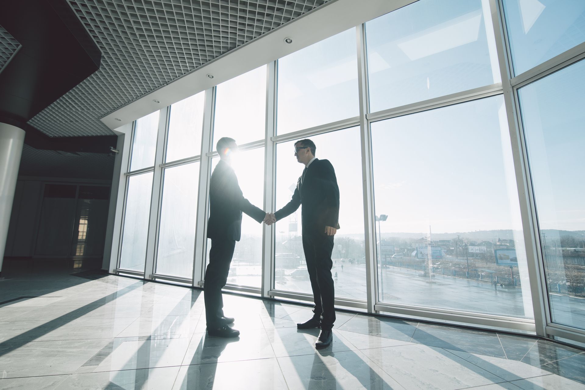 Two businesspeople shaking hands in a modern office with large windows, sunlight, and city views.
