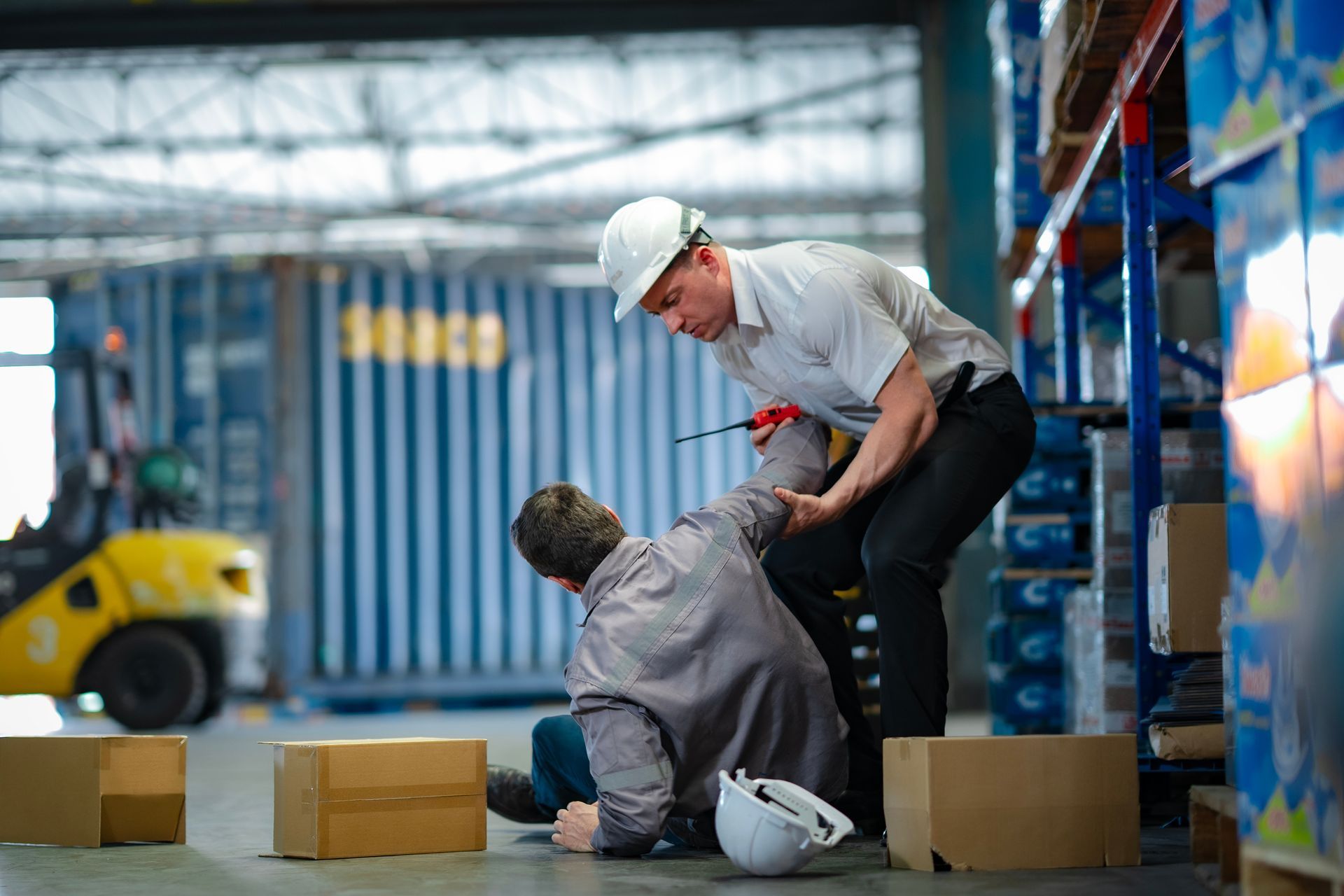 Warehouse worker helping injured colleague after a fall. Boxes and forklift visible. Warehouse worker helping injured colleague after a fall. Boxes and forklift visible.
