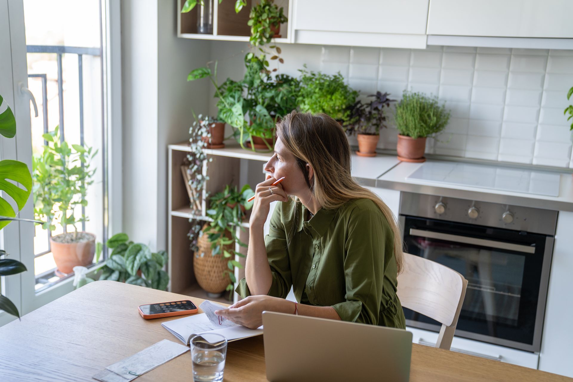 Woman in green shirt, at a table, looks out window, holds papers, a laptop and plants in background. Woman in green shirt, at a table, looks out window, holds papers, a laptop and plants in background.