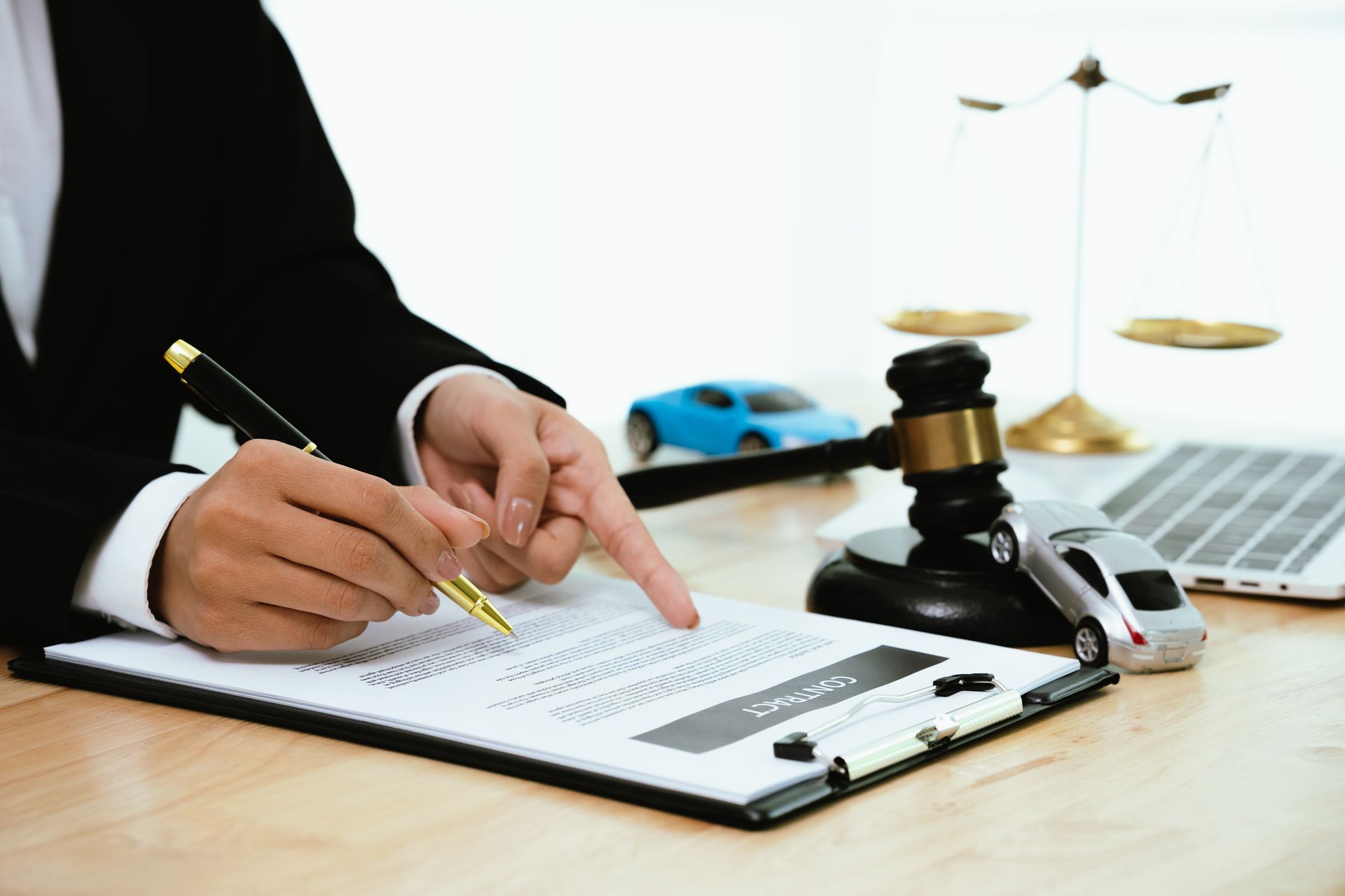 Person in suit pointing at contract on desk with toy cars, scales, and gavel. Person in suit pointing at contract on desk with toy cars, scales, and gavel.