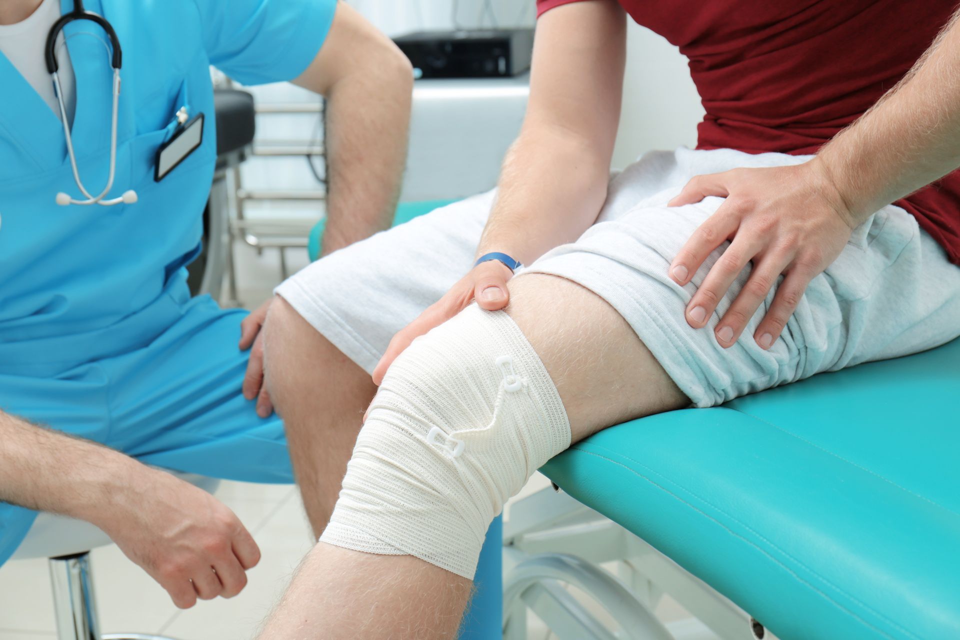 Person with bandaged knee being examined by a healthcare worker in an exam room.