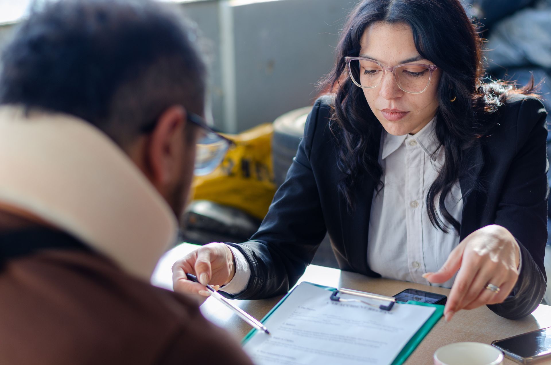 Woman in glasses points at paperwork with a man wearing a neck brace, at a table. Woman in glasses points at paperwork with a man wearing a neck brace, at a table.