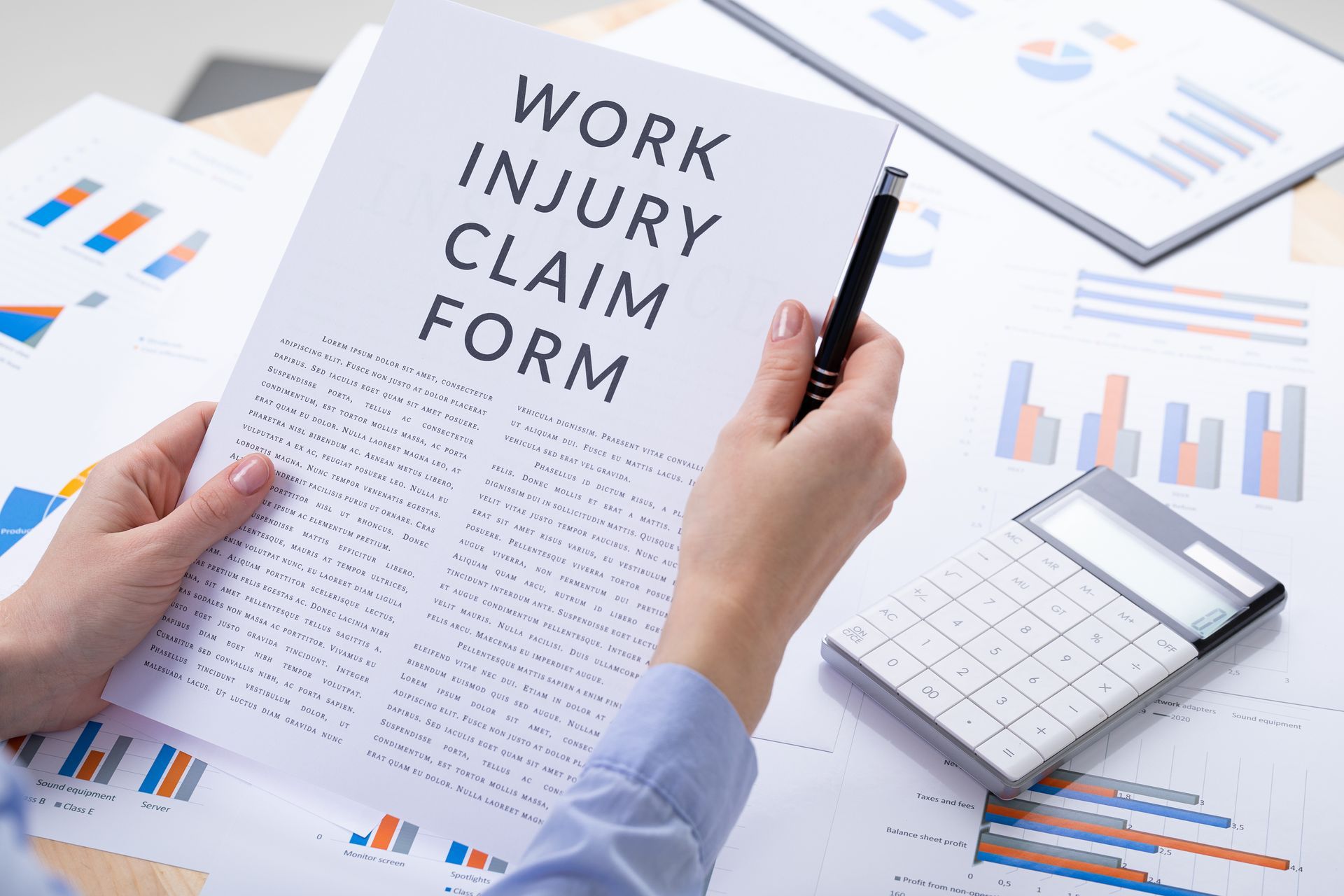 Hands holding a work injury claim form, next to a calculator and paperwork. Hands holding a work injury claim form, next to a calculator and paperwork.