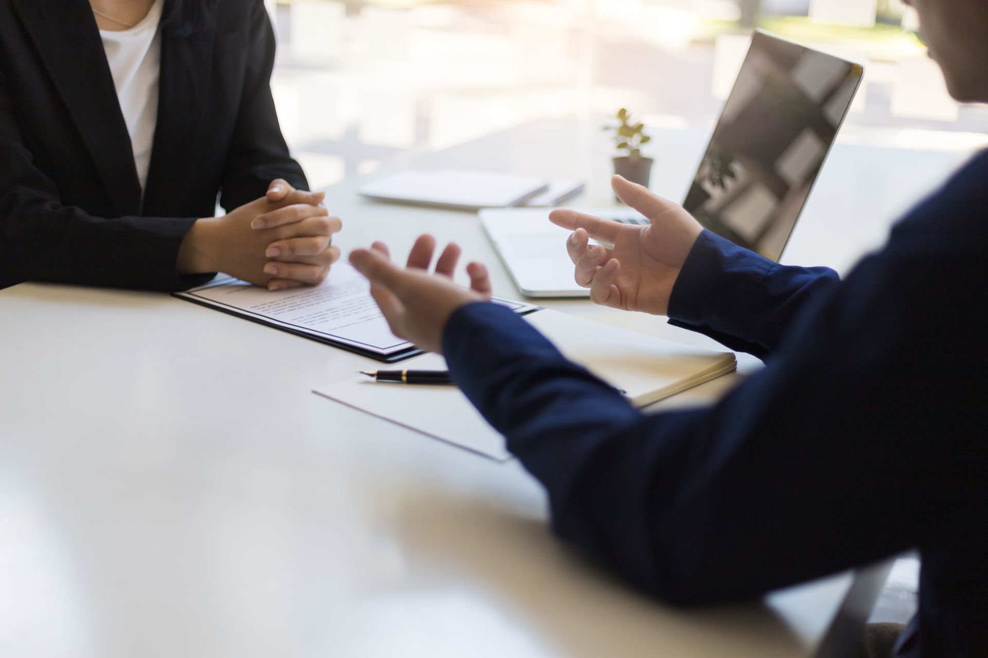Two people in business attire at a table; one speaking, gesturing; the other listening, hands clasped. Two people in business attire at a table; one speaking, gesturing; the other listening, hands clasped.