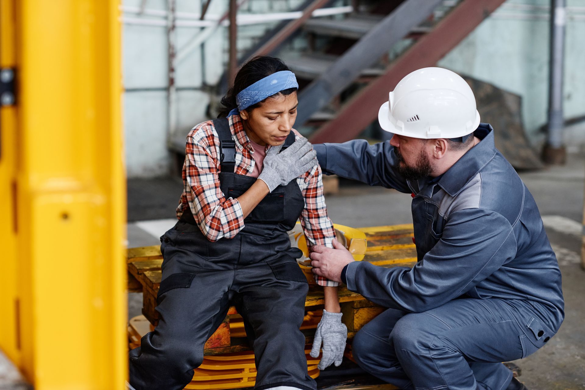 Construction worker assisting a colleague who appears injured. Factory setting with yellow machinery. Construction worker assisting a colleague who appears injured. Factory setting with yellow machinery.