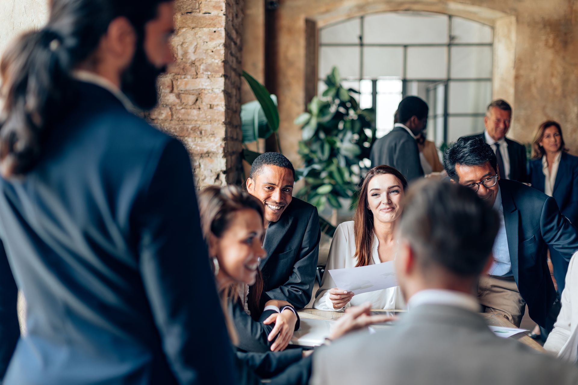 Businesspeople in a meeting, some seated at a table, others standing. Smiling faces, formal attire, in an office space.