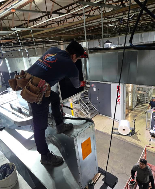 Worker on a metal structure inside an industrial building, wearing a safety harness and tool belt.