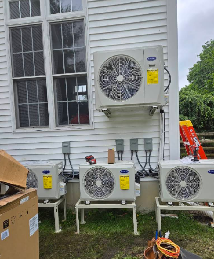 Multiple outdoor HVAC units mounted and on stands beside a white house, with boxes and orange cord on the lawn.