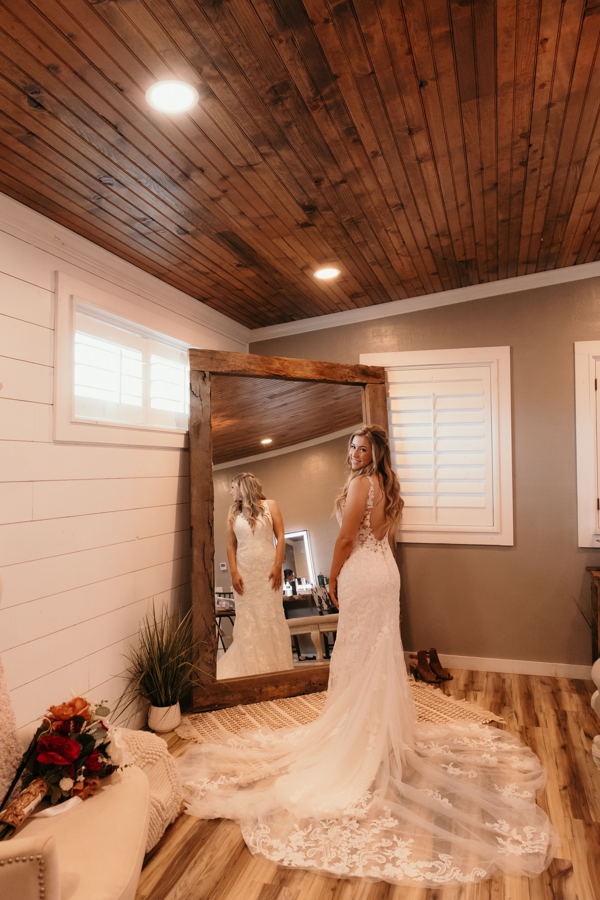 Bride in a cream lace gown standing in a rustic room beside a large mirror and wooden ceiling.