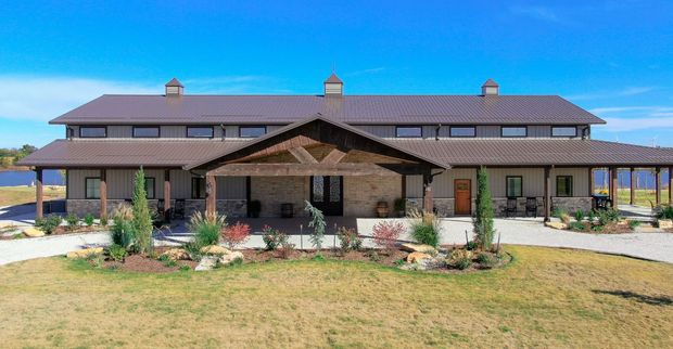 Large rustic lodge with a covered porch and dark roof in a sunny grassy landscape, mountains in the distance
