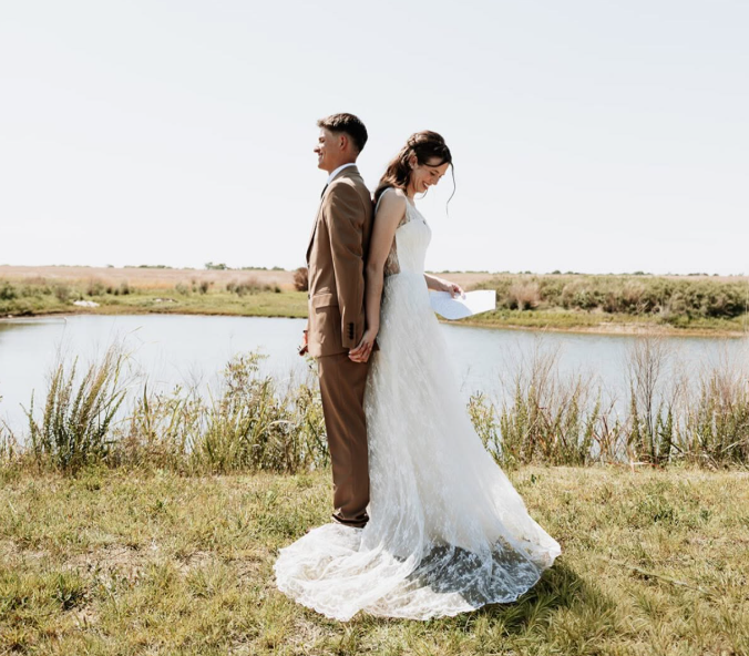 Bride and groom standing back-to-back by a lakeside in a white wedding dress and brown suit