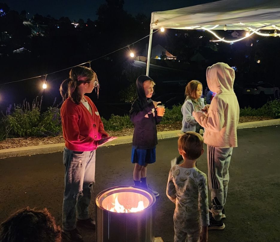 A group of children are standing around a fire pit at night.