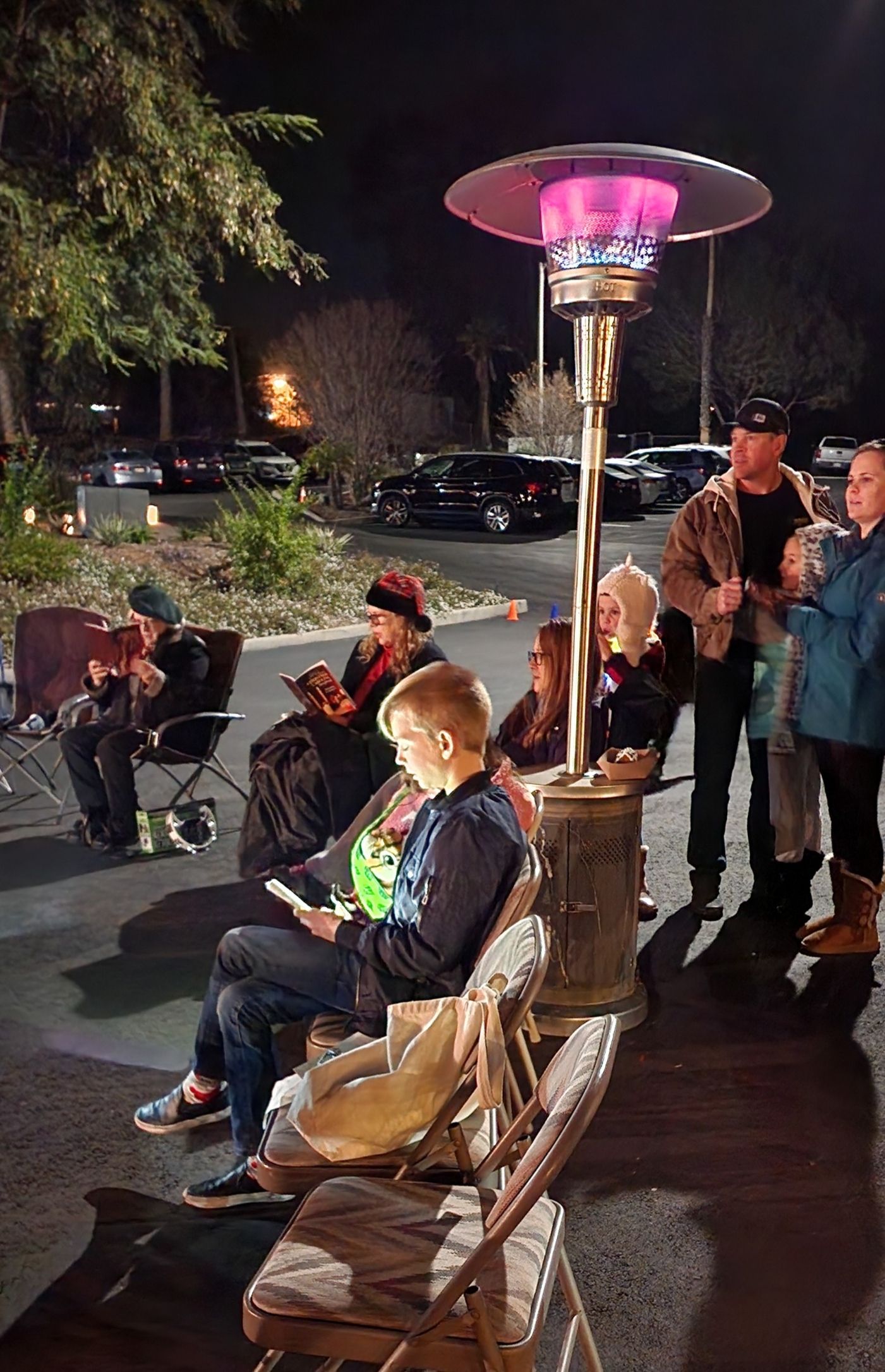 A group of people are sitting under a heater in a parking lot at night.