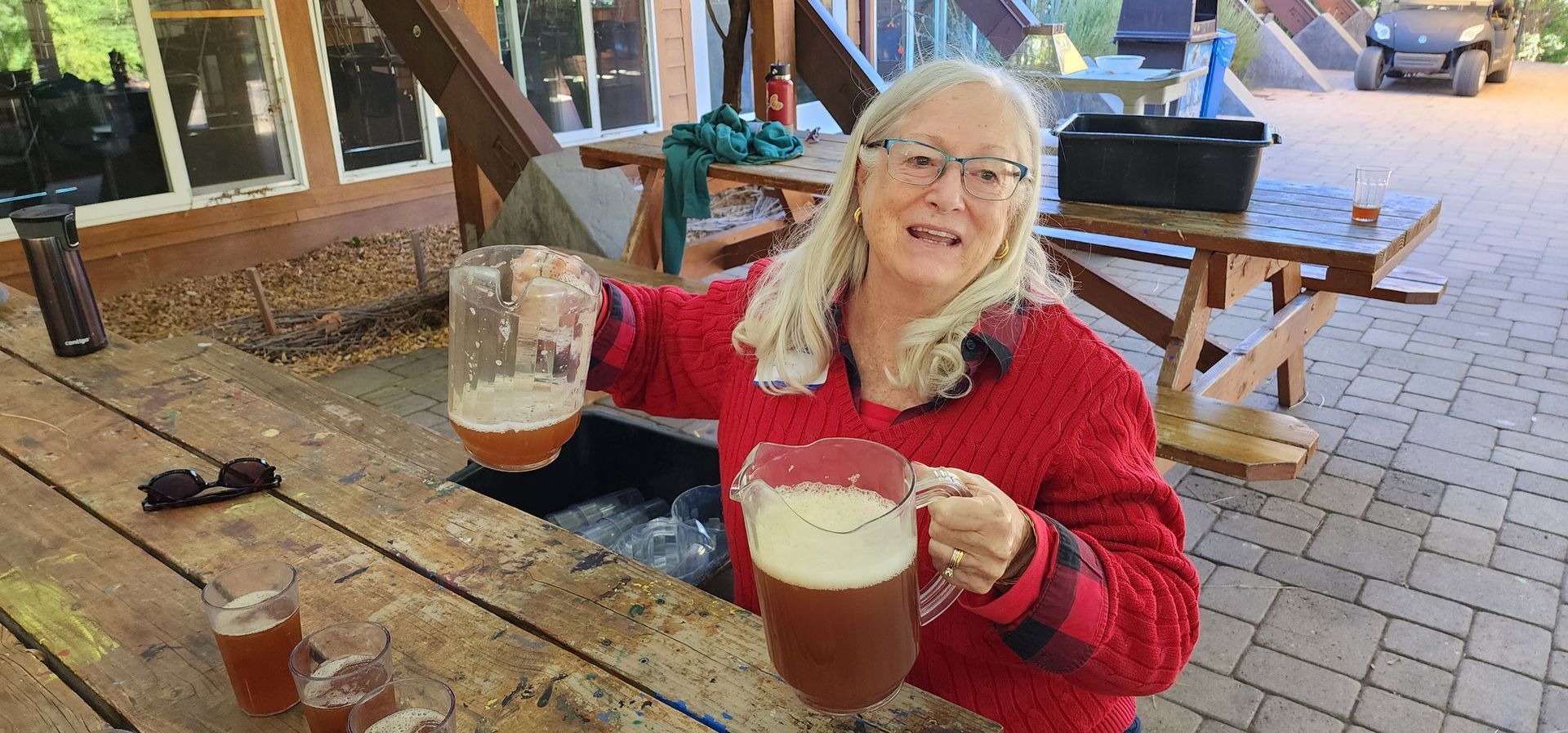 Woman holding a jar and pitcher of beer, smiling. She's outside on a wooden table, with glasses of beer.