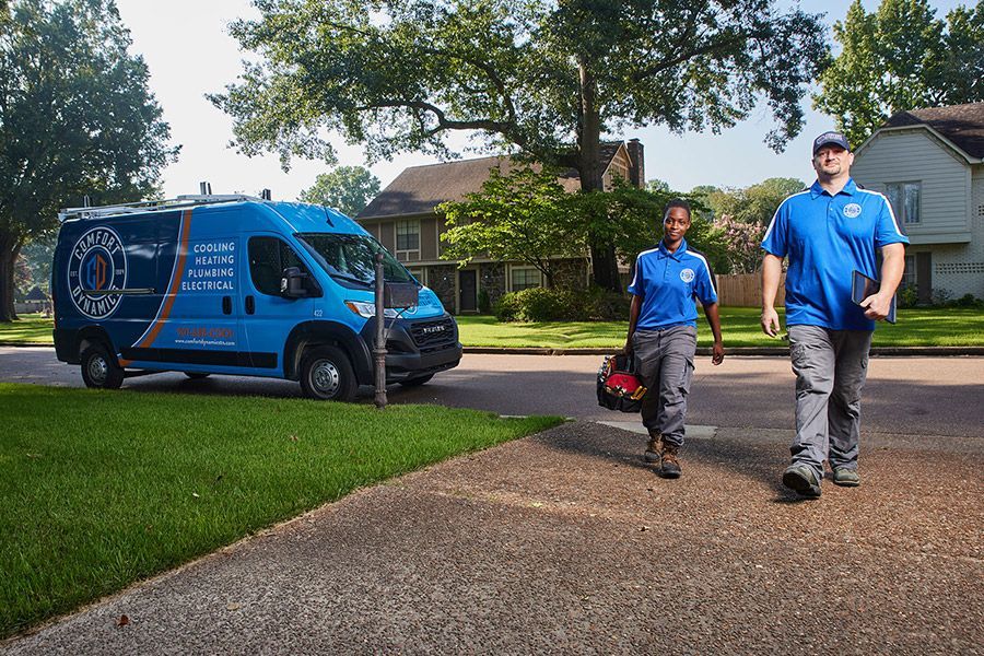 Two service technicians in blue uniforms walk toward a house, van parked on the street.