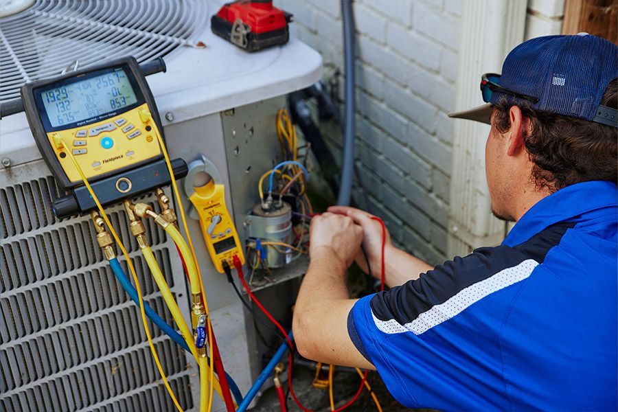 HVAC technician in a blue shirt tests an AC unit's electrical components with tools and gauges.