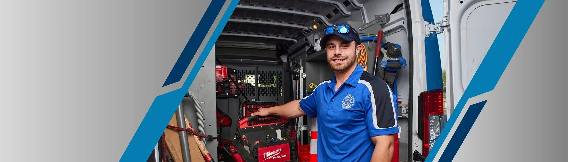 A smiling HVAC technician stands in a van. He wears a blue shirt and cap, gesturing towards his equipment.