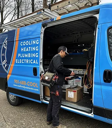 HVAC technician in front of a service van with open doors, blue and orange graphics, outdoors.