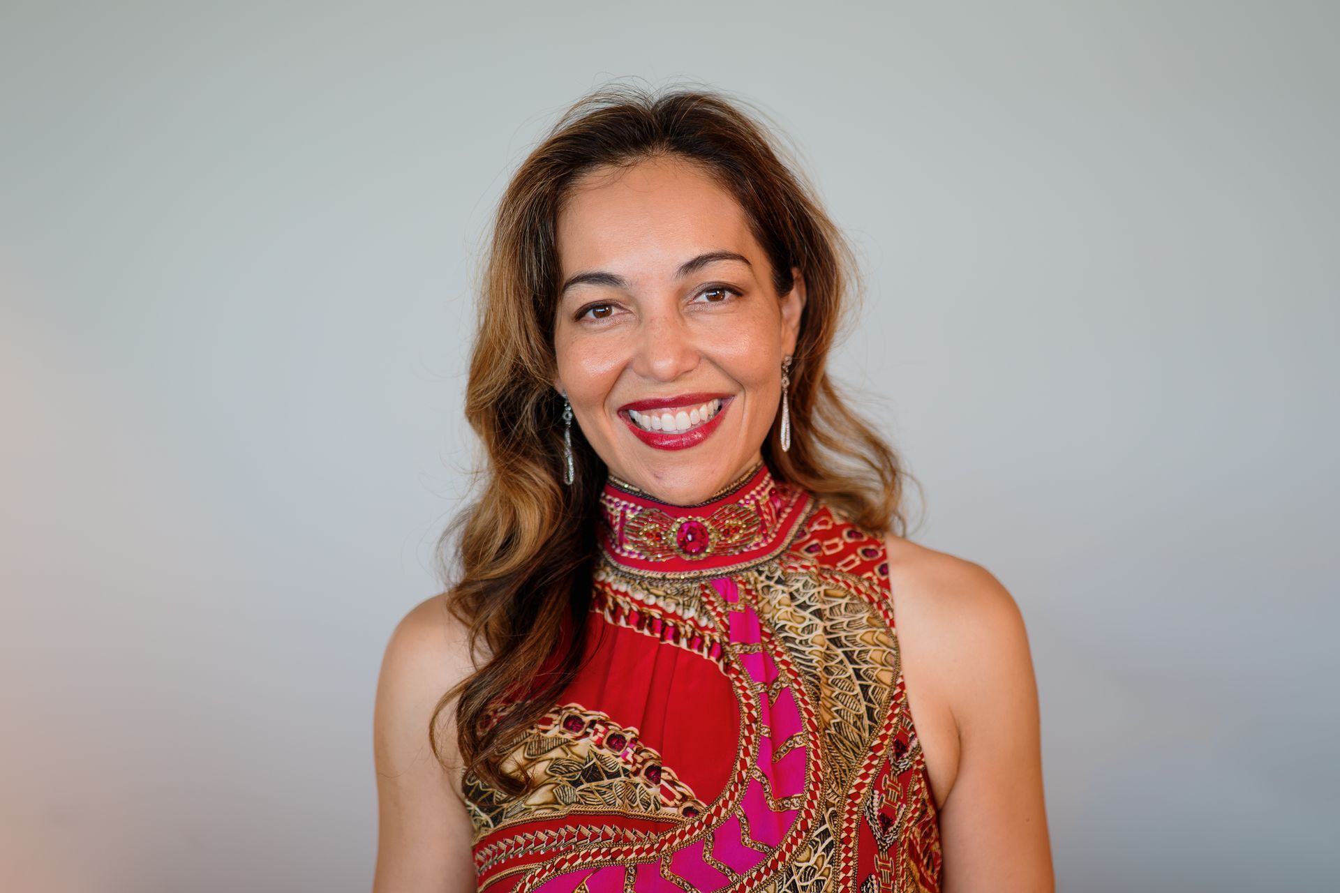 Aurora Bariatrics team member, Associate Professor Ruth Blackham
with long wavy hair, wearing a high-neck patterned dress with ornate detailing, smiling in front of a textured orange and cream tile wall