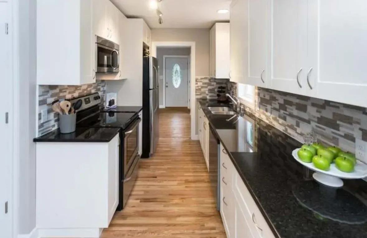 Long, narrow kitchen with white cabinets, black countertops, and wood floor.