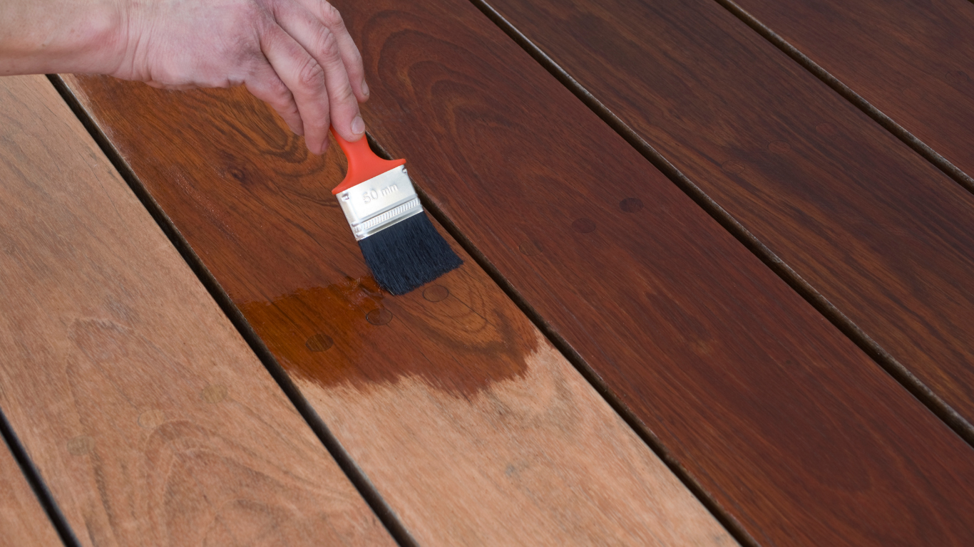 A hand applies wood stain to wooden planks with a paintbrush, creating a contrast between stained and unstained surfaces.