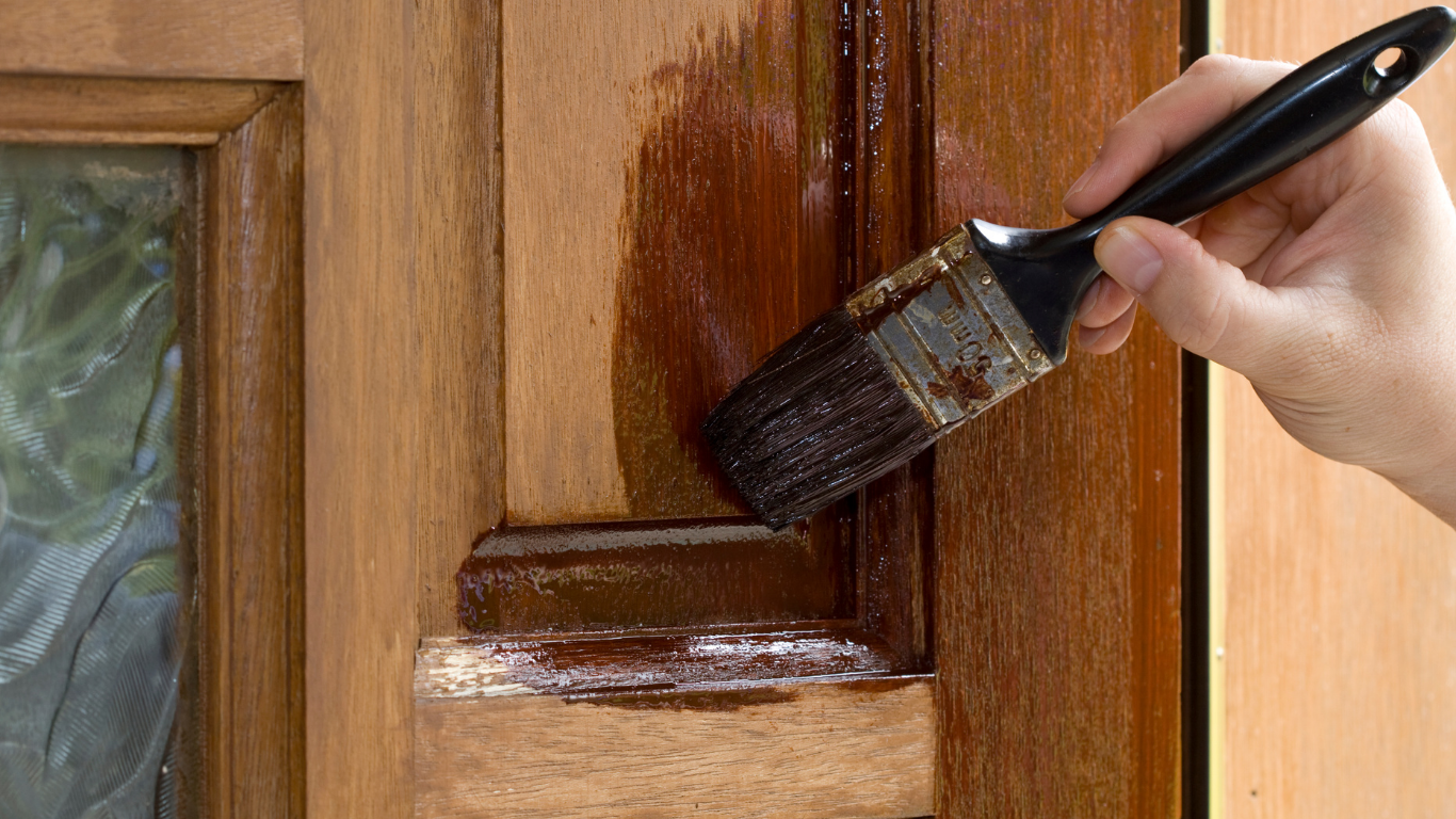 A hand applies dark stain with a brush to wooden trim on a door.
