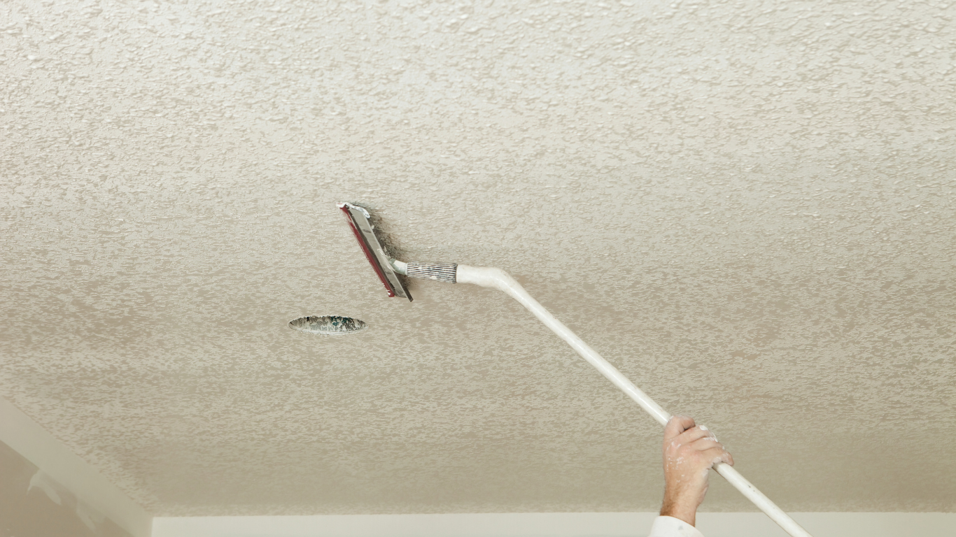 Person scraping a textured ceiling with a long-handled scraper. The ceiling is light-colored with a popcorn texture.