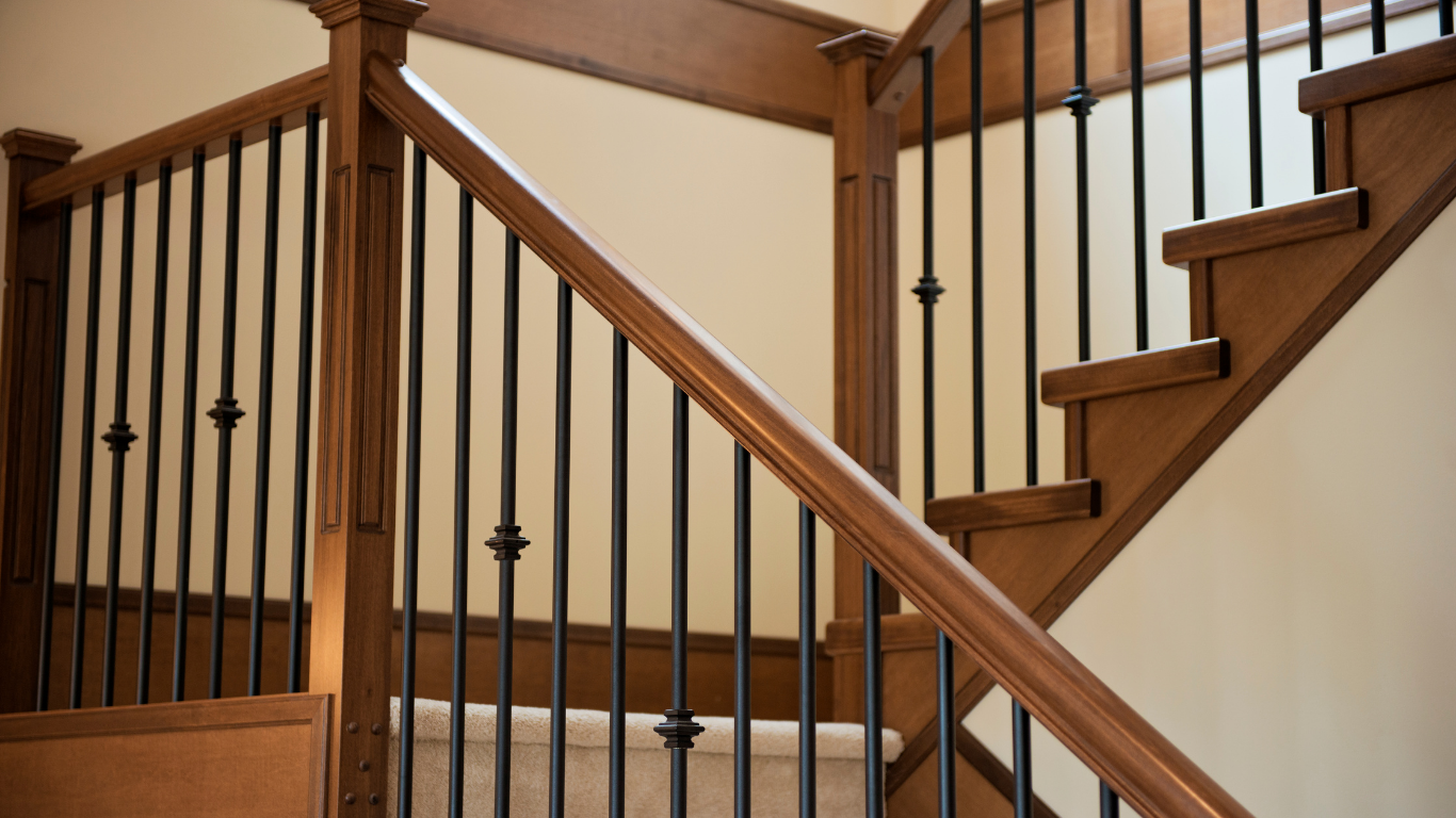 Wooden staircase with black metal balusters and a light-colored carpet runner, set against a neutral wall.