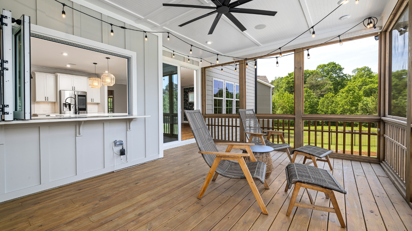 A screened porch with wooden flooring and railing, an outdoor seating area, and a pass-through window to a kitchen.