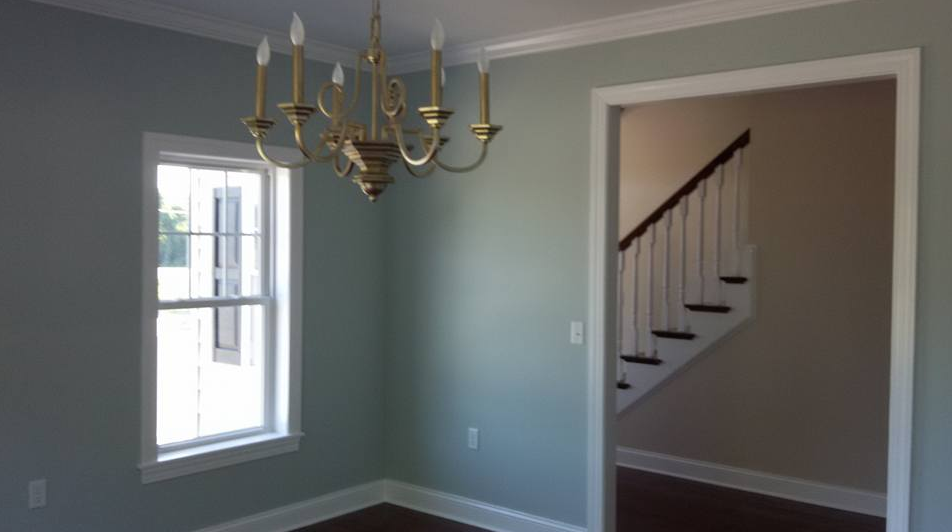 A dining room with pale blue walls, a gold chandelier, a window, and a doorway leading to a staircase.