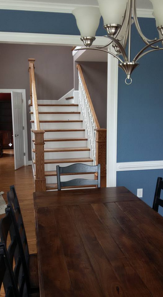 A dining room with a wooden table and stairs. Blue and gray walls with white trim and a chandelier are visible.