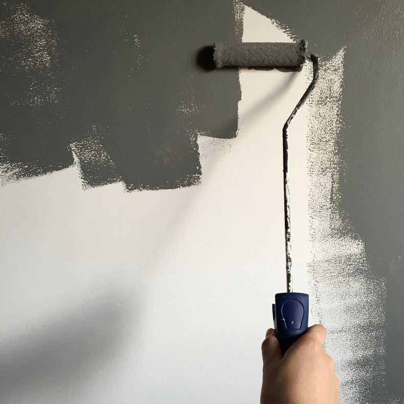 Hand holding a paint roller applying grey paint to a white wall.