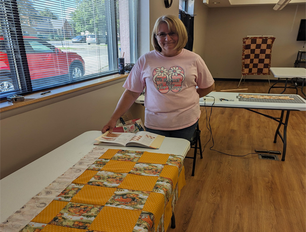 A woman in a pink shirt is standing next to a table with a quilt on it