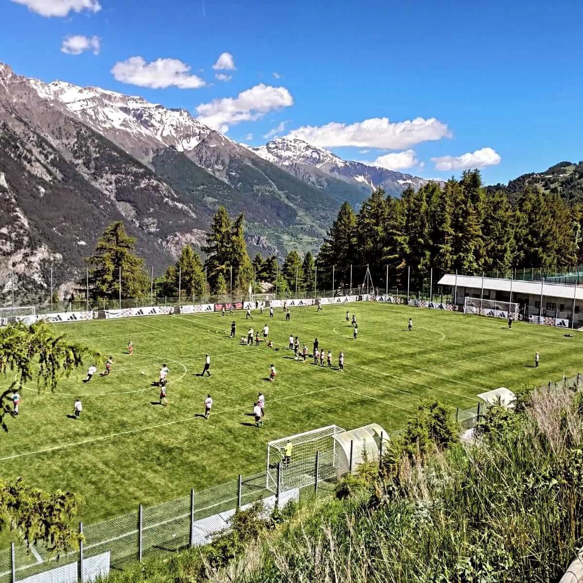 Un gruppo di persone sta giocando a calcio su un campo con le montagne sullo sfondo.