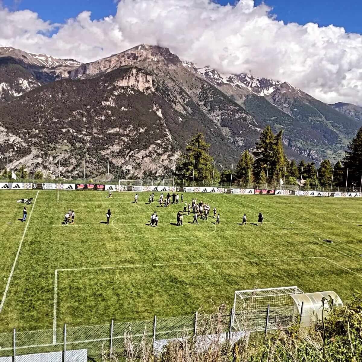 Un gruppo di persone sta giocando a calcio su un campo con le montagne sullo sfondo.
