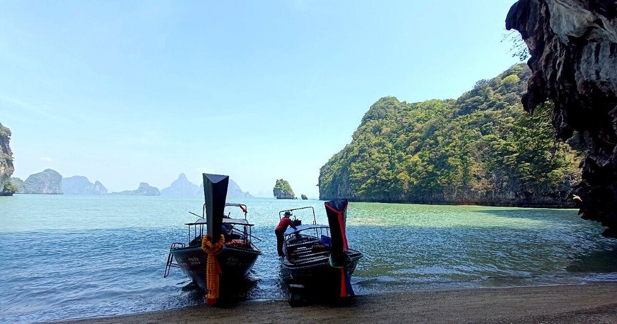 Two longtail boats pulled up on a beach below limestone cliffs in Phang Nga Bay