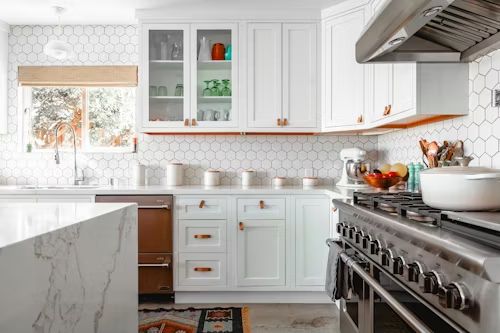 A bright kitchen featuring white cabinets, a marble-topped island, a large stainless steel stove, and hexagon wall tiles.