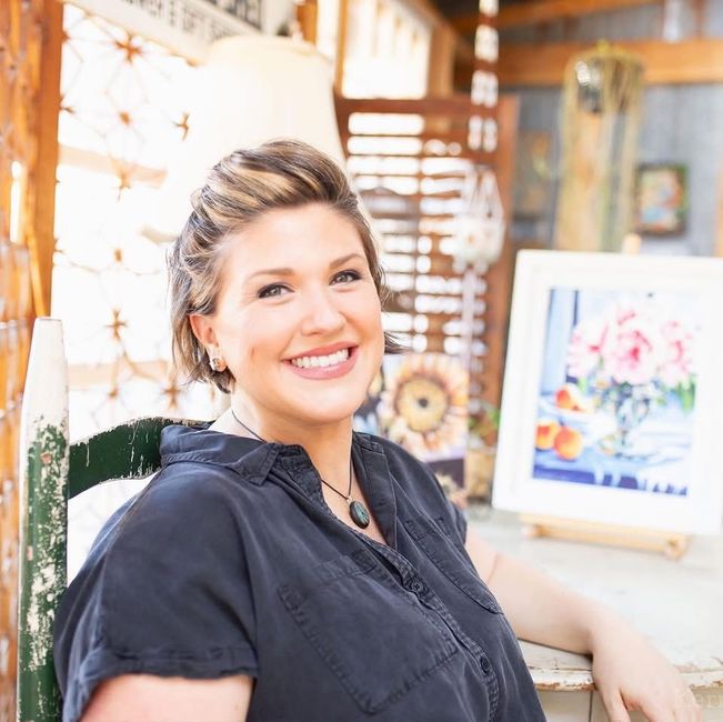 Woman smiling, holding a paintbrush in front of a painting of flowers. She's in a rustic setting, soft light.
