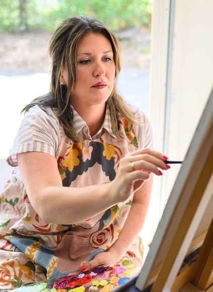 Woman smiling, holding a paintbrush in front of a painting of flowers. She's in a rustic setting, soft light.