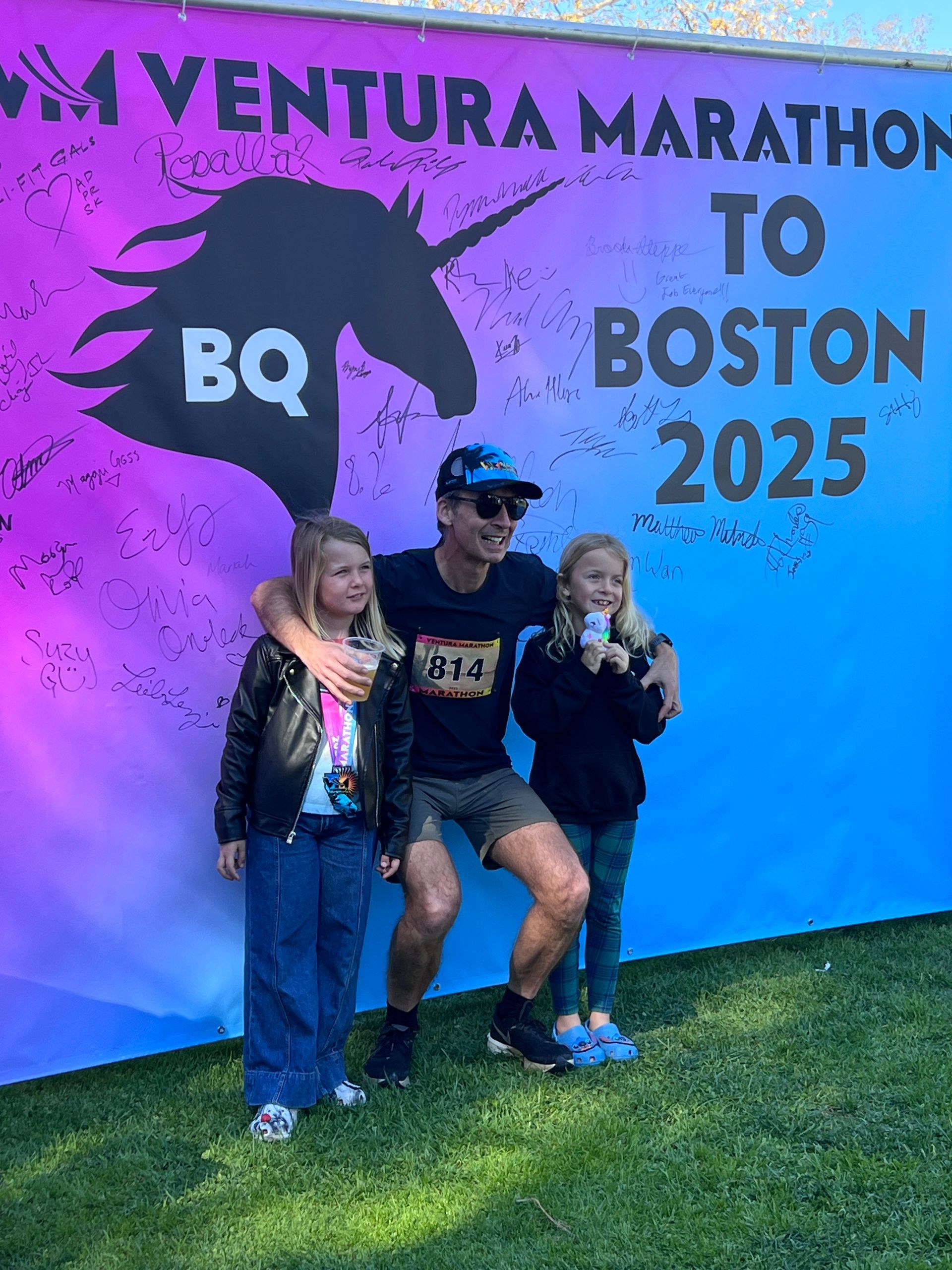 Runner + two kids at the Boston 2025 banner, signed by runners who qualified for the Boston Marathon at the Ventura Marathon.
