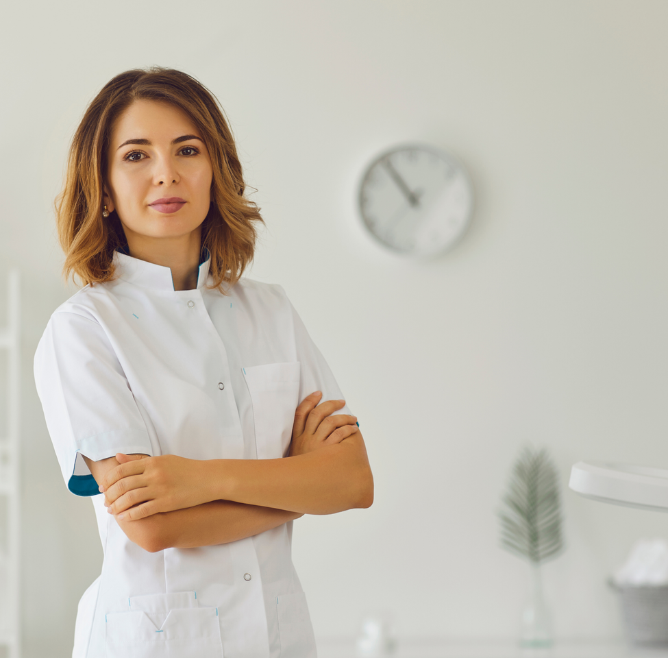 A confident female healthcare worker in a white coat standing in a bright, minimalistic room at a professional footcare centre.