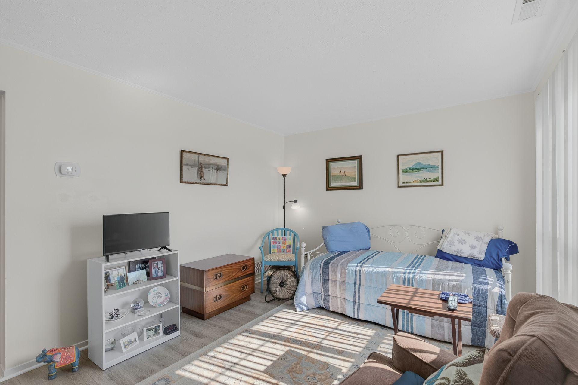 Small bedroom with a blue bed, wooden chest, TV stand, and framed wall art.