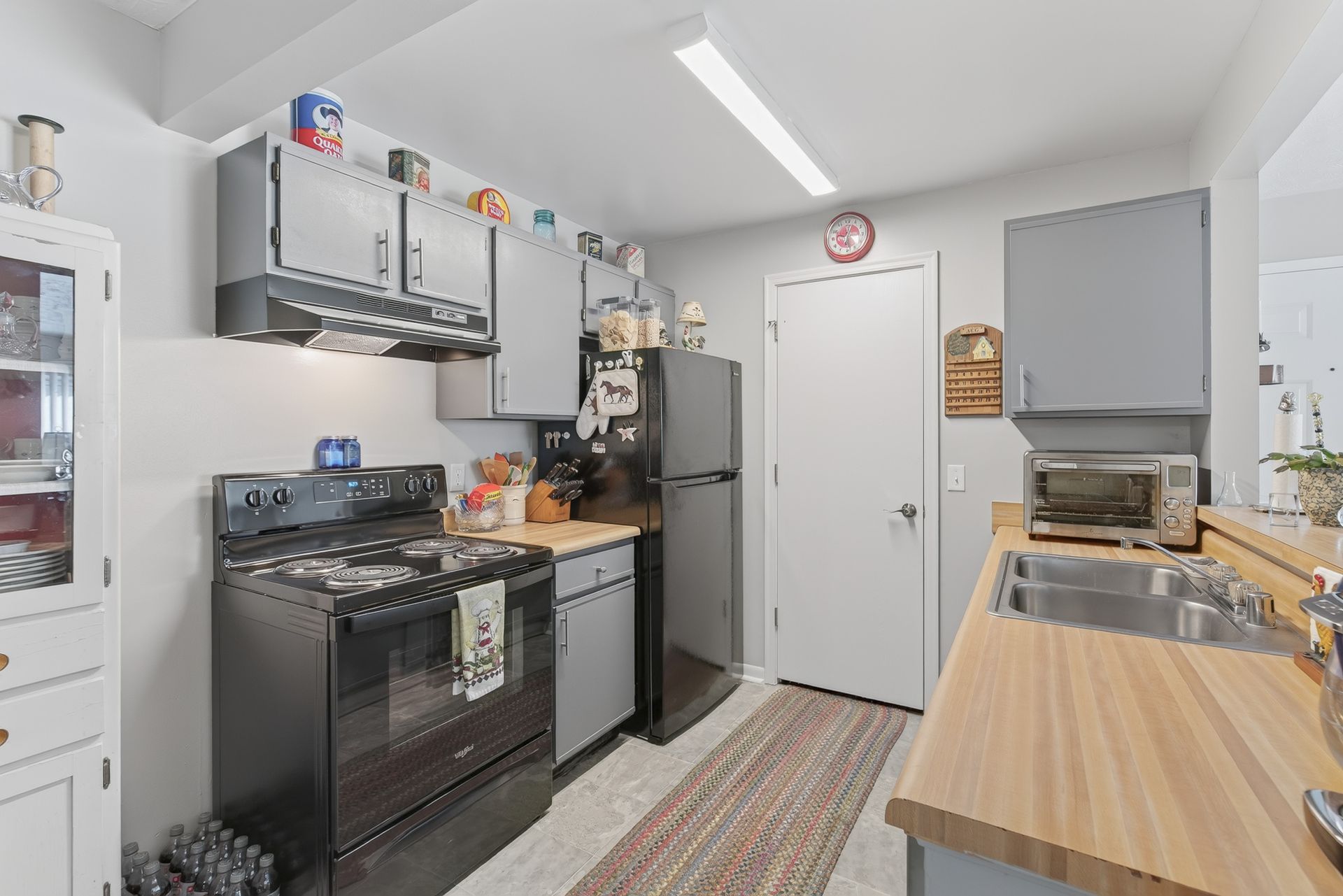 Small kitchen with black stove and fridge, light wood countertops, and gray cabinets near a white door.