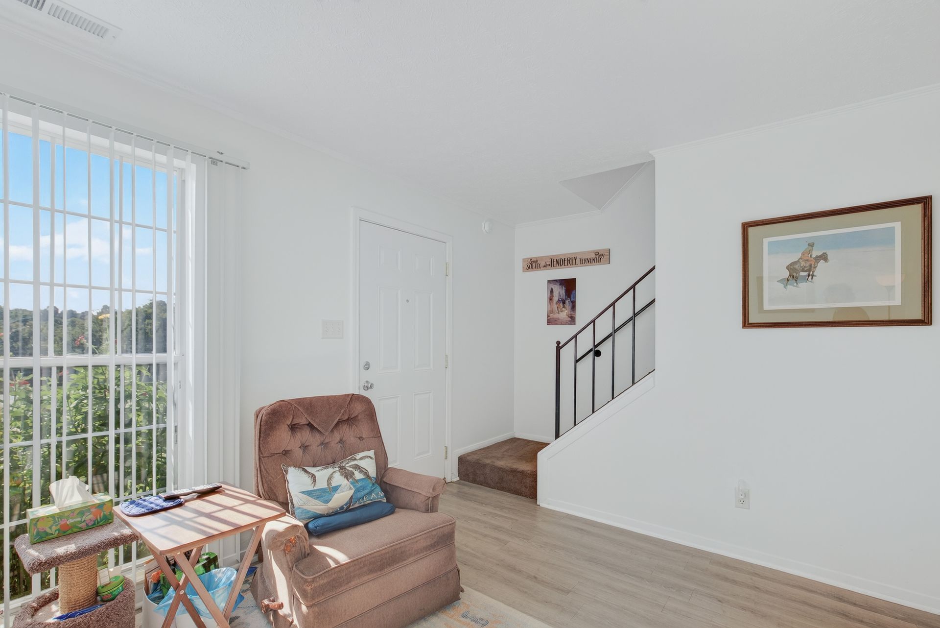 Bright living room with recliner, side table, staircase, and large window with blinds.