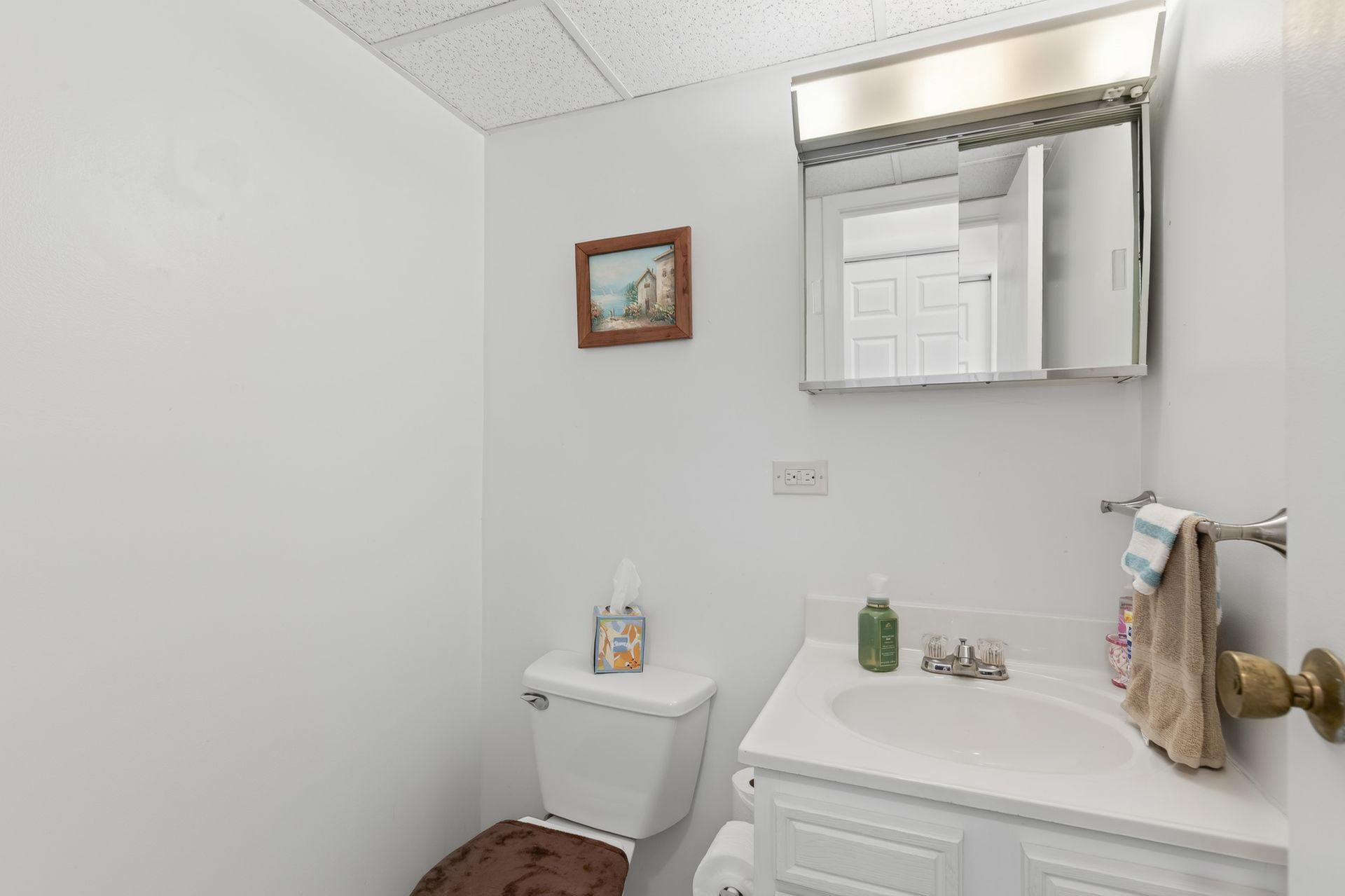 Small white bathroom with sink, toilet, mirror cabinet, and a framed picture on the wall.