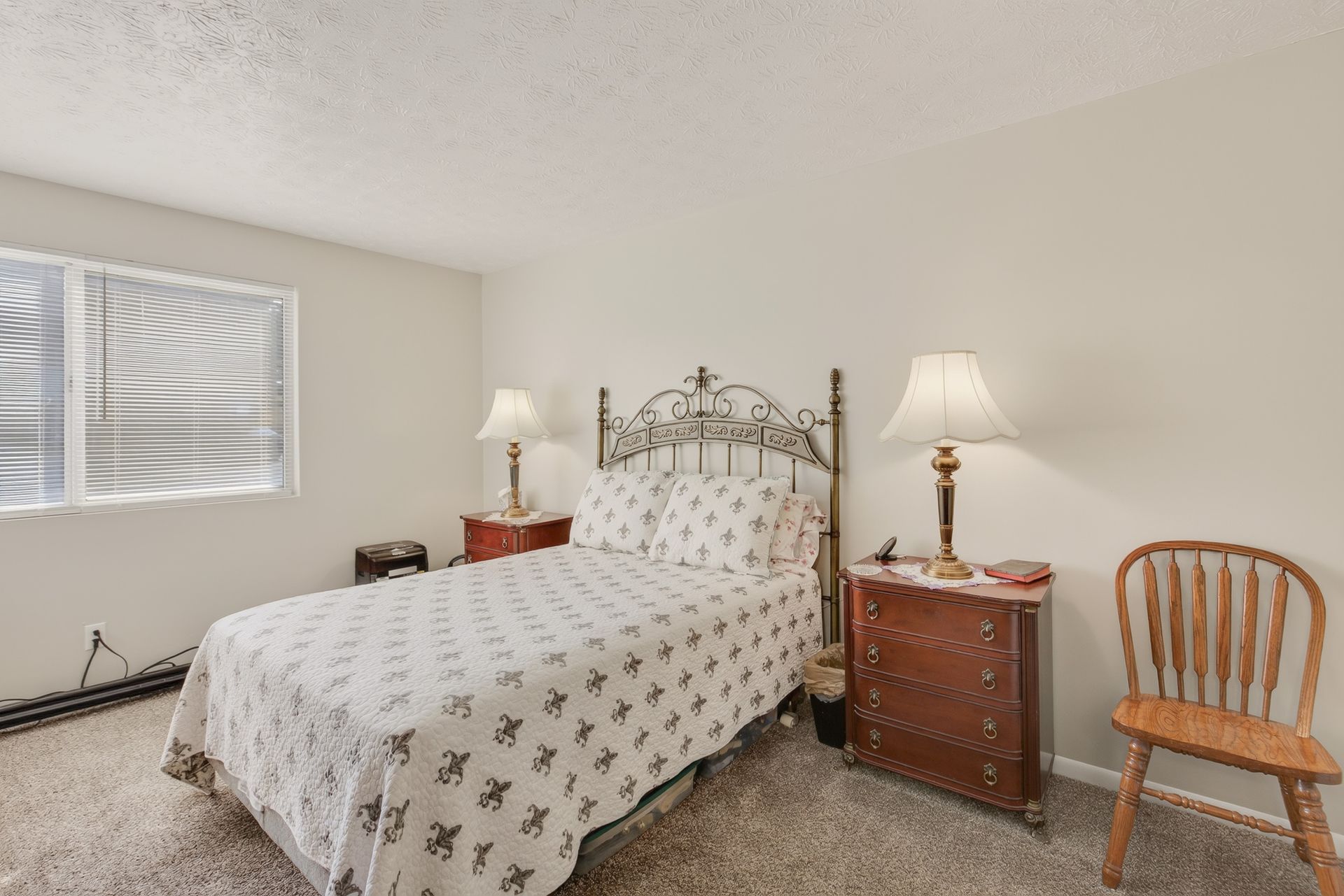 Bedroom with wrought-iron bed, patterned bedding, nightstands, lamps, and a wooden chair by the window
