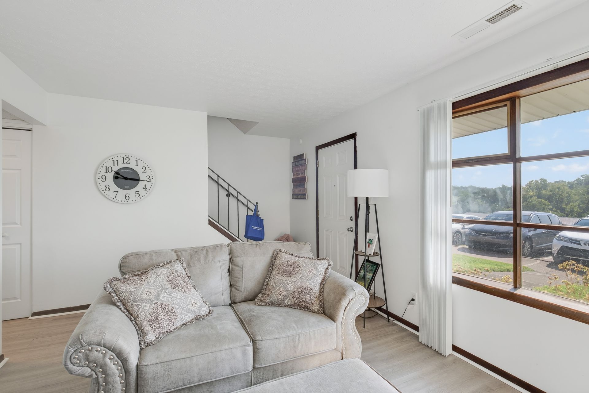Bright living room with a light sofa, wall clock, floor lamp, and large window with a view outside.