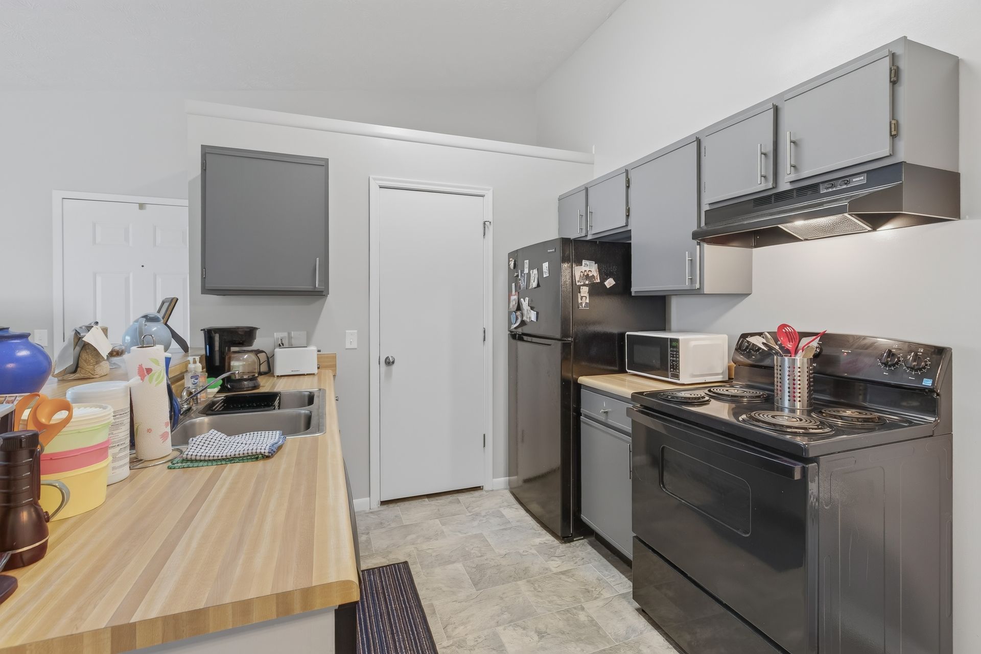 Small kitchen with gray cabinets, black appliances, and a cluttered counter beside a wooden table.