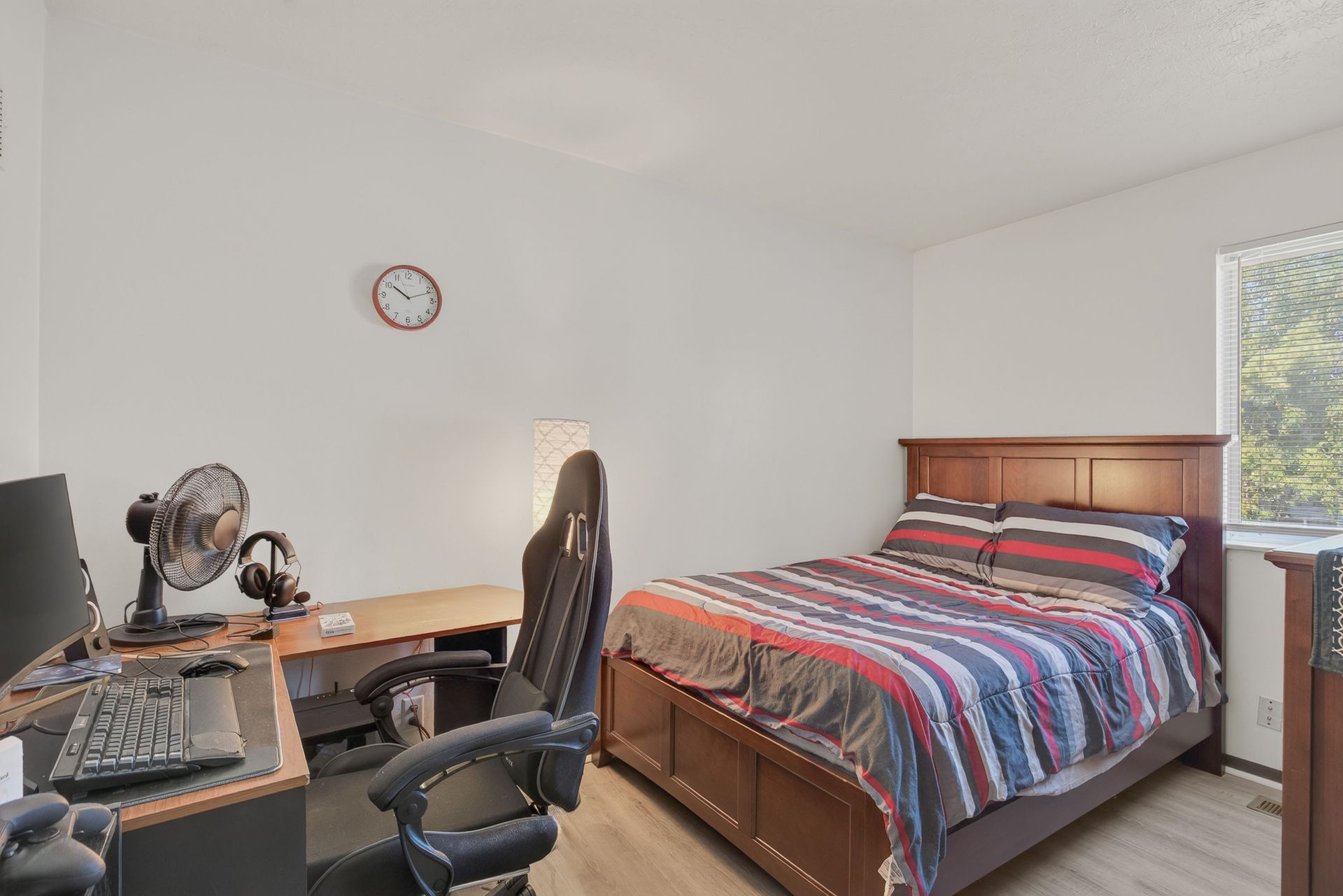 Bedroom with a bed, desk, computer chair, and fan beneath a wall clock.